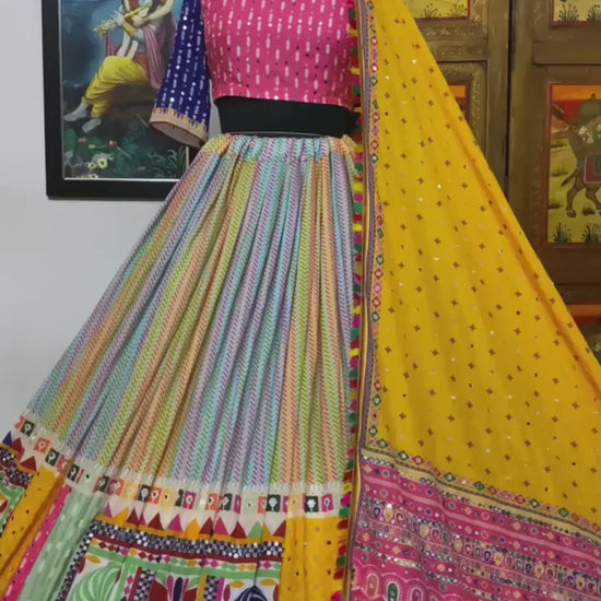 A beautifully designed Gujarati lehenga displayed on a mannequin, featuring a multicolored skirt with detailed patchwork, a pink and blue embroidered blouse, and a bright yellow dupatta with traditional mirror work and embellishments. A painting of Radha-Krishna is visible in the background.







