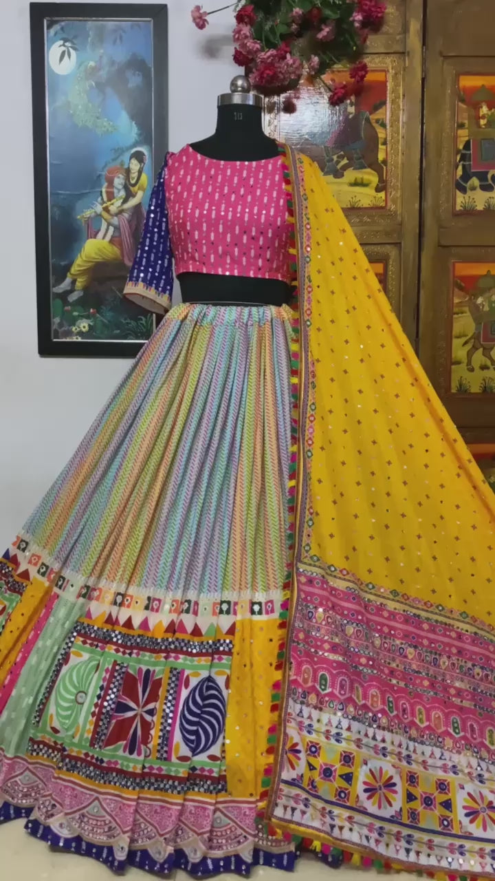 A beautifully designed Gujarati lehenga displayed on a mannequin, featuring a multicolored skirt with detailed patchwork, a pink and blue embroidered blouse, and a bright yellow dupatta with traditional mirror work and embellishments. A painting of Radha-Krishna is visible in the background.







