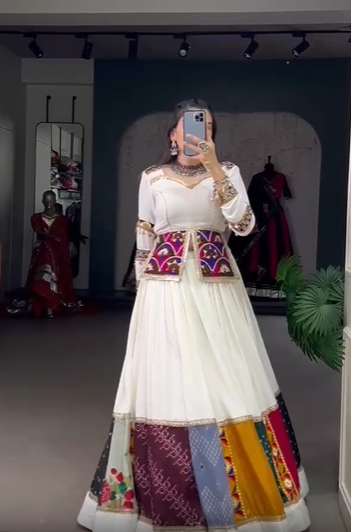 A woman wearing a traditional Gujarati lehenga admires herself in the mirror, showcasing the outfit from different angles. The lehenga features a white base with vibrant patchwork panels, intricate embroidery, and mirror work. She gracefully twirls and adjusts the ensemble, highlighting its elegant design in a boutique setting.







