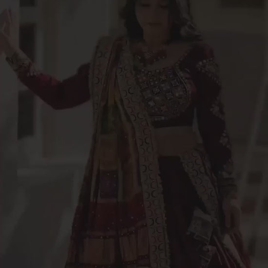 A woman dressed in a stunning maroon designer lehenga gracefully swirls, showcasing the outfit's elegant 4-meter flair. The lehenga features intricate mirror work, a canvas patta border, and vibrant traditional prints. As she twirls, the rich fabric and detailed craftsmanship are highlighted, making it a perfect attire for festive occasions like Navratri and weddings.
