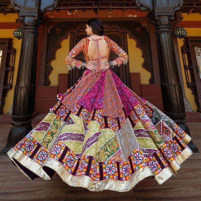 A woman gracefully twirls in a vibrant Gujarati lehenga choli, showcasing intricate embroidery, mirror work, and a backless blouse with tie-up details. The colorful patterns and flowing silhouette create a mesmerizing visual against a heritage-style architectural backdrop.